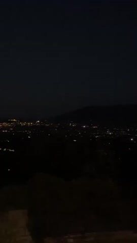 The video shows a nighttime view of a city spread out across a valley. The city lights are a prominent feature, appearing as numerous small, glowing dots. In the background, mountains rise against the dark sky. The foreground is darker, with the silhouettes of trees and bushes obscuring a wall or barrier. The overall impression is one of a panoramic vista of a city at night.