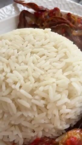 The video features a close-up shot of a prepared meal served on a white plate. On the left side of the plate, there is a neatly mounded portion of fluffy white rice. To the right of the rice, a rich, reddish stew is visible. As the camera slowly pans across the dish, more details of the stew become apparent.

The stew appears to be tomato-based, with visible pieces of red crab, sliced onions, and what look like dark-colored mushrooms or other vegetables. The crab pieces are reddish-orange, suggesting they have been cooked into the stew. The overall presentation is that of a ready-to-eat dish, likely a local meal from Iwofe, Nigeria, given the typical ingredients and presentation. The setting appears to be indoors, likely a dining table. No people or text are visible in the video.