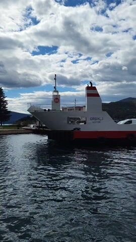 The video shows a stationary ferry named "GRBALJ KOTOR" at a dock on a body of water. The ferry is painted white with red accents. A few vehicles are visible on the ferry. In the background are mountains and a cloudy sky with patches of blue. A tree is visible on the left side of the frame. The water has small ripples, reflecting the sky.