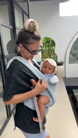 The video shows a woman with her hair in a bun, wearing sunglasses and a dark t-shirt, carrying a baby in a blue carrier. The baby is wearing a white bonnet and a striped shirt. The woman is smiling and looking at the baby, who is looking back at the camera. They are standing outdoors in what appears to be a modern, minimalist setting with plants visible in the background. The woman adjusts the baby's carrier strap. The baby waves its hand. The woman makes a kissing face. The baby looks at the camera with a serious expression. The woman speaks.