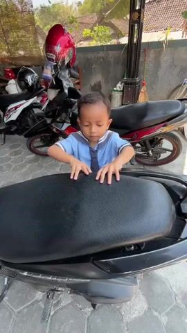 A young boy is reaching into the storage compartment of a black scooter. The scooter seat is open and a plastic container of food, containing rice, green beans, and possibly chicken, is visible inside. The boy is wearing a light blue shirt with white stripes on the collar and sleeves. In the background, several other motorcycles are parked, and a building with a tiled roof is visible. The ground is paved with interlocking gray stones.