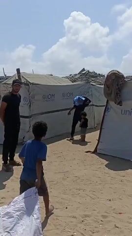 A wide shot captures an outdoor scene with several tents set up on sandy ground. Two children are walking away from the camera down a narrow path between the tents. The child in the foreground is wearing a blue t-shirt and shorts, and is pulling a large white bundle on the ground. The child behind them is wearing a black shirt and shorts. Further down the path, a person in a dark shirt and blue shorts carries a blue object on their head. In the background, a pile of debris and damaged structures are visible under a bright, cloudy sky. The tents are white and bear logos including "IOM UN MIGRATION" and "UNICEF". The overall environment appears to be a makeshift settlement.