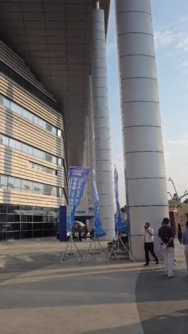The video shows a wide shot of an outdoor area next to a modern building with large, cylindrical columns. Several people are walking away from the camera towards a display of flags and some vehicles. The flags are various colors and appear to be from different organizations. The vehicles seem to be part of an exhibition or trade show. The sky is blue with some clouds, and the lighting suggests it is daytime. There are also some promotional banners and stands visible. The overall atmosphere appears to be that of a public event or gathering.