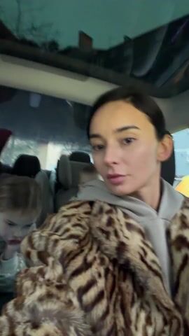 The video shows a woman in the front seat of a car taking a selfie video. She is wearing a grey hoodie and a fur coat with a leopard print pattern. Behind her are two young children in car seats. One child is looking at the camera, and the other is looking down. The car is parked on a street with buildings visible through the windows.