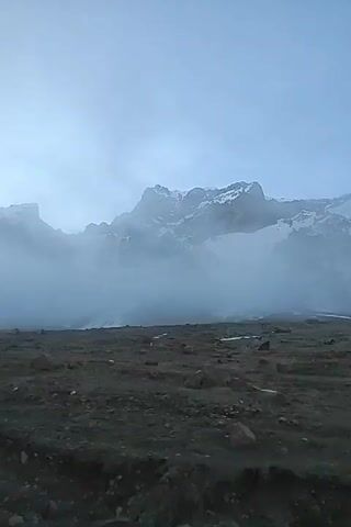 A sweeping shot captures a vast, desolate landscape under a cloudy or misty sky in what appears to be Kanobi, Georgia. Snow-capped mountains are partially obscured by dense fog or low-hanging clouds in the background, creating a dramatic and somewhat mysterious atmosphere. The foreground consists of barren, rocky, and uneven terrain, suggesting a high-altitude or rugged environment. No people or prominent objects are visible, emphasizing the raw, untouched nature of the scene. The lighting is soft and diffused, indicating either early morning, late afternoon, or heavily overcast conditions. The camera slowly pans or moves across the landscape, offering a wide view of the expansive, unpopulated area. No text is visible in the video.