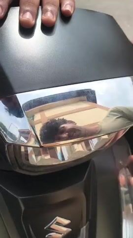 Two pairs of hands are visible on a black scooter. One pair, on the left, grips the front of the scooter. The other pair, on the right, is also touching the front of the scooter. A reflection in a chrome accent on the scooter shows the face of a person with curly dark hair looking upwards at a building. The building is tan with brown trim and blue windows. The sky is visible and appears to be daytime.