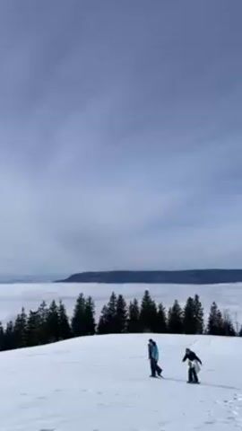 The video shows a snow-covered field on what appears to be a mountain top. The sky is a light blue with patchy clouds. Several pine trees are visible, and a layer of fog or clouds fills the valley below. Several people are moving around on the snow-covered field. The camera pans across the landscape showing more of the snow-covered area, trees, and cloud cover. There are trails in the snow, possibly from skis or snowshoes.