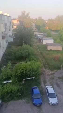 The video shows an aerial view of a residential area, likely during the early morning or late afternoon given the lighting. The scene captures several apartment buildings, parked cars, and overgrown vegetation. In the foreground, a large apartment building with multiple balconies dominates the left side of the frame. Below it, two cars are parked on a patch of ground surrounded by greenery. Further into the scene, a narrow, unpaved road runs between more apartment buildings and rows of garages. The road continues into the distance, where it is lined with trees and houses, suggesting a more rural or suburban setting beyond the immediate residential complex. Several utility poles and wires are visible throughout the video, indicating the presence of infrastructure. The overall impression is one of a somewhat unkempt but lived-in urban or semi-urban environment.