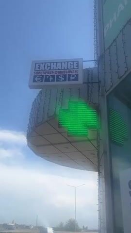The video shows an exterior view of a modern building under a bright, sunny sky with some clouds. A prominent sign above the building reads "EXCHANGE" in English and "ՏԱՐԱԴՐԱՄԻ ՓՈԽԱՆԱԿՈՒՄ" (Money Exchange) in Armenian. Below this text, symbols for the Euro (€), Pound (£), Dollar ($), and Ruble (₽) are displayed.

Attached to the side of the building is a large, blinking green LED cross, which is a common symbol for a pharmacy. Partially visible text on the building's facade to the right includes "PHA" (likely part of "PHARMACY") and "ST".

In the foreground, a multi-lane road with light traffic is visible. A dark-colored car drives past from left to right. A street light pole can be seen in the distance, and the horizon reveals more structures or an open landscape. The building itself has a curved, somewhat futuristic design with external lighting fixtures. The time of day appears to be daytime, with clear visibility and bright illumination. The context provided specifies the location as Yerevan, Armenia, which is consistent with the Armenian text on the sign.