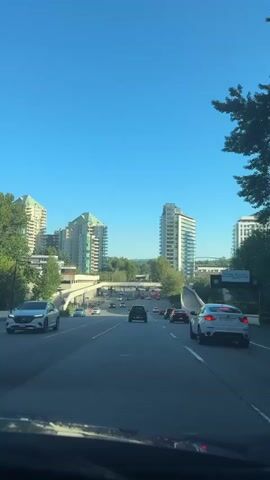The video shows a view from the driver's seat of a car on a highway. The sky is clear and blue. On the left side of the highway are several modern buildings, with green roofs on some of them. On the right side of the highway, there is a concrete barrier and some trees. There are many cars on the highway, moving in the same direction. The car is moving forward at a steady pace. The traffic appears to be light to moderate. The overall impression is of a sunny day in a city with modern architecture.