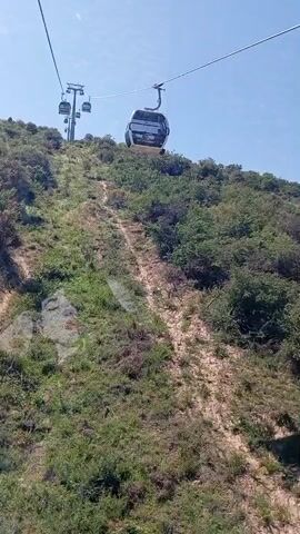 A view from a cable car ascending a hill. Another cable car passes by heading in the opposite direction. The hillside is covered in green vegetation and a dirt path is visible. The sky is blue. A person is visible in the passing cable car.