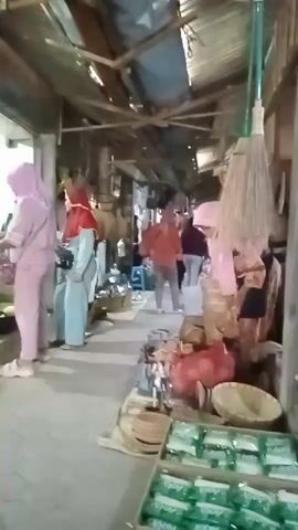 This video shows a market scene. Several people are walking and browsing through stalls. The stalls appear to be selling various goods, including woven baskets, household items, and what looks like packaged food products. The market has a rustic feel, with a corrugated metal roof and exposed wooden beams. The lighting suggests it might be daytime, possibly overcast. The people are dressed in casual clothing, with some individuals wearing hijabs. The overall atmosphere seems to be one of a typical local market.
