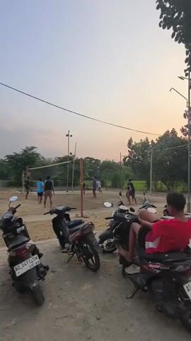 The video shows an outdoor volleyball game at dusk. Several people are playing volleyball on a sand court with a net. There are also several motorcycles parked on the side of the court. A few people are sitting on the motorcycles watching the game. The sky is clear with a few clouds. The lighting suggests it is either early morning or late evening.