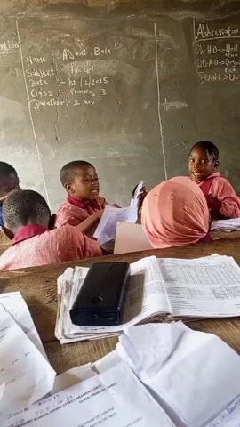 The video shows several young children in a classroom setting. The students, wearing red school uniforms, are seated at wooden desks, with papers and books scattered around. A chalkboard behind them contains handwritten text, including the title "Abbreviation & Meaning" and a list of abbreviations like WHO, OAU, and UN. One child is holding up a piece of paper and appears to be speaking or reciting something. The classroom is simple and somewhat rustic, with natural light coming in through a window. In the foreground, a black power bank rests on one of the desks. The overall impression is that of a primary school lesson in progress.