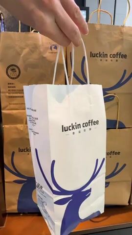 A hand holds a white paper bag with a blue deer logo and "luckin coffee" printed on it. The bag is in front of several larger brown paper bags, also with the "luckin coffee" logo. The video then switches to a lime green paper bag decorated with black polka dots and various cartoon images, including a pineapple, a reindeer character, a coffee cup, a palm tree, sunglasses, a flamingo, and a peace sign. The bag also has "CROCS LUCKIN" and a heart icon printed on it. The green bag is held in front of the same background of brown "luckin coffee" bags. Finally, the video pans up to show the interior of a store, with a circular blue and white "luckin coffee" sign visible, as well as more "luckin coffee" bags.