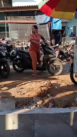 A woman in an orange jumpsuit is sitting on a dark colored scooter in an outdoor setting that appears to be a market or a busy street. She is looking to her right and appears to be waiting. There are other scooters parked around her and some stalls or shops in the background with colorful awnings. The ground is dirt and some construction materials are visible in the foreground. The woman is standing with her right foot on the ground. She is wearing sandals. She has long dark hair. She is holding the handlebars of the scooter. The scooter has a dark colored body and silver accents. The scooters in the background are a mix of colors. There are people in the background, some of whom are walking or standing near stalls. The overall lighting suggests it is daytime.