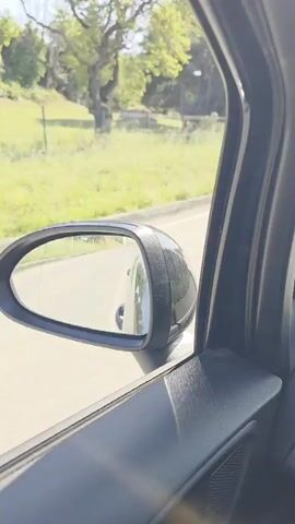 The video presents a view from inside a moving vehicle, looking out the passenger side window. The car's black side mirror is visible in the lower left quadrant, reflecting a portion of the vehicle's side. Outside the car, a light-colored paved road is present, alongside a grassy verge. Beyond the verge, a dark metal fence runs parallel to the road, enclosing a green, slightly elevated area dotted with numerous coniferous trees. A light pole and a small white marker post are visible along the roadside. The scene is brightly illuminated by sunlight under a clear sky. Audible speech in a foreign language, possibly Italian, accompanies the visuals.