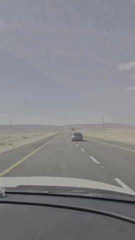 A car is driving on a long, straight, paved desert highway. There are two lanes going in each direction, separated by a broken white line. The sky is slightly overcast, and there is a shadow on the top left corner of the windshield. In the distance, a sign is visible, along with power lines. There is another car ahead of the driver.