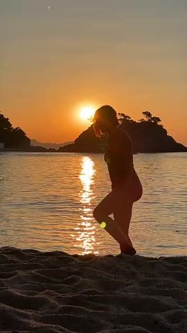 A person in a bathing suit is silhouetted against a sunset on a beach. The sun is low on the horizon, casting a warm orange glow over the water. The person is walking along the shoreline, with waves gently lapping at their feet. The sand is dark and textured in the foreground. In the background, there is a rocky island with trees. The person is in motion, with their arms and legs extended.