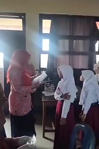 A group of students in hijabs and school uniforms stand in a classroom. A teacher holds a piece of paper and appears to be speaking to the students. Some students are looking at the teacher, while others are looking away or at each other. The room has desks and chairs, and windows with blinds. The lighting suggests it is daytime.