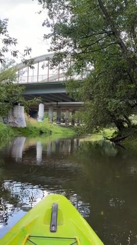 The video is shot from the perspective of someone in a bright yellow kayak moving down a calm river. The kayak's bow is in the foreground. On the left side of the river, there is a concrete bridge support with graffiti on it. The bridge itself is visible above, with a pedestrian walkway or a bike path running across it. The bridge has a curved arch structure. Trees line the right bank of the river, with lush green foliage. The water reflects the bridge and the trees, creating a mirrored image. The sky is overcast, with visible clouds. The overall impression is a peaceful paddle through a natural setting with a piece of urban infrastructure nearby.