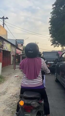 A person wearing a purple headscarf, a striped long-sleeved shirt with purple trim, and black pants is riding a motorcycle in traffic. The motorcycle is a dark color, possibly black or gray, with a visible "HONDA" logo on the back. The rider is wearing a black helmet. Behind the rider, another person is partially visible, also wearing a helmet. The motorcycle is in a lane with several cars. To the left of the road, there are small shops or storefronts with red doors and a sign for "VINA LAUNDRY". The road surface appears to be paved with some gravel and dirt. The sky is cloudy with a mix of blue and gray. The overall scene depicts a typical traffic situation in an urban or suburban area.