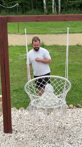 A man is standing in front of a white net swing hanging from a wooden frame. He is wearing a white t-shirt and black shorts. The swing is on a gravel ground with grass and trees in the background. The man bends over to interact with something inside the swing, then straightens up with a smile. He bends over again, reaching into the swing, and then straightens up again.