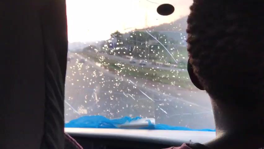The video shows a person sitting in a vehicle looking out the window. The window is covered in raindrops and has a crack. Outside the window, there is a road and some greenery. The person's head is silhouetted against the bright light outside. The vehicle appears to be moving. A blue piece of material is affixed to the bottom of the window.