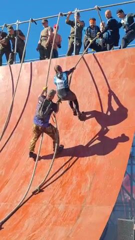 In this vertical video, two individuals are ascending a steep, curved orange wall using ropes, as part of an obstacle course. The person in the foreground, wearing a purple backpack and camouflage pants, is climbing a thick rope with their hands and feet. Above and to the right, another person in athletic wear is also using a rope to climb. They are both casting prominent shadows on the wall. Several people are positioned at the top of the wall, holding onto a horizontal bar and ropes, appearing to assist or supervise. The sky is a clear, bright blue. The overall scene depicts a challenging physical activity in an outdoor setting.