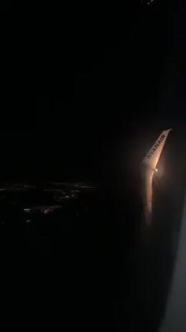 The video shows a view from an airplane window at night. The wing of a Ryanair plane is visible, with a light on the tip of the wing. Below, a city's lights are faintly visible in the distance. The rest of the scene is dark.