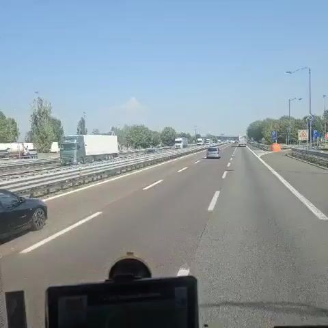 The video shows a view from inside a vehicle traveling on a multi-lane highway on a clear, sunny day. The highway is flanked by green trees and vegetation. Other vehicles, including cars and trucks, are visible on the road. The video appears to be recorded by a dashboard camera or similar device, as a portion of the vehicle's interior, including a screen or mount, is visible at the bottom of the frame. The highway has white lane markings and a metal guardrail separates the lanes of traffic from the opposite direction and the shoulder. There are signs and streetlights along the highway. The camera perspective is from the driver's seat, looking forward.