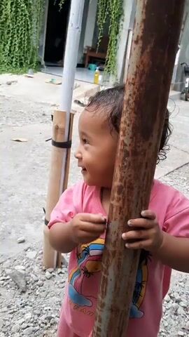 A toddler with dark hair and a pink t-shirt is smiling and holding onto a rusty metal pole. The toddler's right hand is touching the pole near the chest. The toddler's left hand is also on the pole, with fingers wrapped around it. The background shows a building with green vines hanging down, a white post, and some gravel on the ground. The lighting suggests it is daytime.