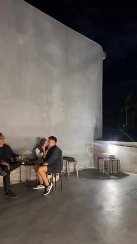 The media file depicts an outdoor evening scene at what appears to be a cafe or restaurant in Tamansari, Indonesia. The time of day is night, indicated by the dark sky and the presence of a full moon. The weather appears clear, with clouds visible around the moon.

The setting is a rooftop or elevated patio area with concrete walls and flooring. Several small tables and stools are arranged on the patio, some lit by decorative strip lighting along the wall. In the background, silhouetted trees and structures suggest an urban or semi-urban environment.

The primary subjects are people gathered at the tables. On the left, a couple sits close together at a table, appearing to be engaged in conversation or sharing a moment. Further to the right, another individual sits alone at a table. In the distance, a third person is visible sitting on a stool. The overall mood seems relaxed and intimate, with the lighting and quiet atmosphere suggesting a peaceful evening gathering. There is no visible text in the media file.