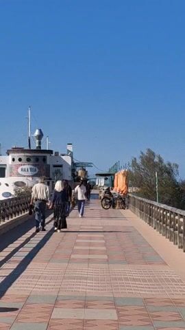 The video shows a brick walkway on a sunny day. Several people are walking along the walkway. There is a building to the left of the walkway that is white with a blue sign that says "MALA VITA." There is a bicycle-powered food cart on the right side of the walkway. A wooden railing borders the walkway. The sky is clear and blue.