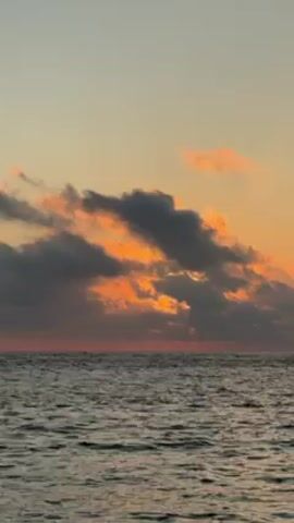 The video depicts a calm ocean scene under a colorful sunset sky. The water has slight ripples and reflections of the sky's colors. Above the horizon, there are layers of clouds; the lower clouds are dark, while the ones above are illuminated with orange and pink hues from the setting sun. The sky transitions from light orange near the horizon to a pale blue at the top of the frame.