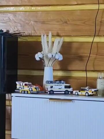 The camera slowly zooms out and pans upward to show three Lego models displayed on top of a white, modern-style cabinet with gold-colored handles. The Lego models are a yellow car with a trailer, a white camper van, and a yellow sports car. Behind the models is a vase filled with artificial cotton and dried pampas grass. A log cabin wall forms the background. A small candle in a glass jar and a gray wireless speaker are also visible on the cabinet. A black TV is mounted on the wall above the cabinet. A black wire hangs down the wall next to the cabinet. The scene is indoors.