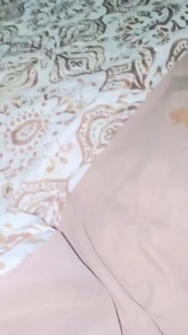 The video shows a close-up view of an orange tabby kitten with a patterned blanket in the background. The kitten is lying on a brown pillow, and a remote control is visible on the pillow near the kitten. The video is shaky and out of focus in some parts, suggesting it was filmed with a mobile device. The kitten appears to be grooming itself or resting.