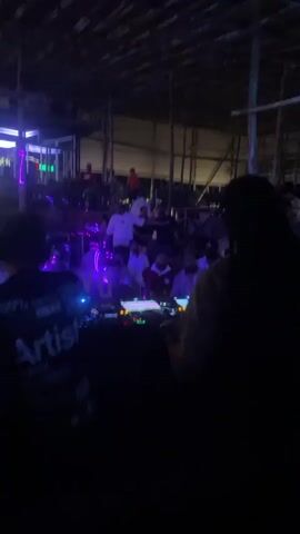 A nighttime scene unfolds within a dimly lit, open-air structure, possibly a bar or club, with a thatched roof and visible wooden beams and supports. In the foreground, two individuals are positioned behind a DJ setup, which includes turntables and a mixer. The person on the left, facing away from the camera, is wearing a dark t-shirt with white text that reads "100% Artist." The person on the right, also facing away, has long dark hair and is wearing a light-colored top. Further back, a crowd of people are visible, some dancing or standing, illuminated by ambient and occasional colorful lights, particularly blue and purple. In the middle ground, a person in a white shirt is holding up a mobile phone with a bright light, possibly taking a photo or recording. Another individual, wearing a patterned shirt, stands to the right of the DJ booth, gesturing with their hand. The overall atmosphere suggests a musical performance or gathering.
