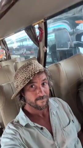The video shows two people on a bus. The first person, a man with graying hair and a beard, is wearing a light-colored, striped button-down shirt and a straw hat. He is in the foreground, looking at the camera and talking. Behind him, a woman with blonde hair is visible. She is wearing a green jacket and a green baseball cap. She is looking off to the side and has her hand near her face. Other passengers and buses are visible through the windows of the bus, suggesting they are at a bus station or a busy thoroughfare. The lighting is natural, likely from the daylight outside.