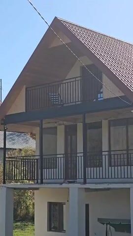 The video shows a two-story house with a brown metal roof. The top floor has a balcony with a black railing and a wooden chair on it. The lower floor also has a balcony with a black railing. The house is surrounded by trees and the sky is blue.