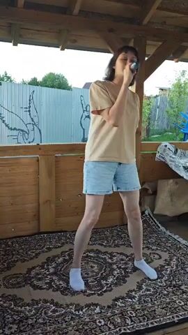 A woman stands on a patterned rug under a wooden canopy. She is wearing a tan t-shirt and light blue denim shorts. She holds a microphone to her mouth and appears to be singing or speaking. Her feet are bare, wearing white socks. The background shows a metal fence with graffiti art, wooden planks, and some foliage.