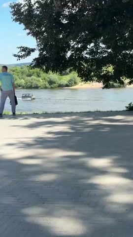 A wide shot shows a paved pathway in the foreground with dappled sunlight. A group of people are visible at different distances. In the mid-ground, on the left, a woman with blonde hair wearing a white top and green pants walks away from the camera. To her right, a man wearing a teal shirt and grey pants stands with his back to the camera, holding a black bag. Further in the background, a river flows with boats visible on it. Trees and greenery line the riverbanks, and hills are visible in the distance under a blue sky with scattered clouds. A statue of a person in a long robe stands on a tiered stone base to the left of the mid-ground. Another man wearing a light-colored shirt and shorts walks away from the camera towards the right. Further to the left and back, a couple stands close together, and another person pushes a stroller. In the far left background, a single lamppost stands near two park benches, one of which is occupied by a person using a laptop.