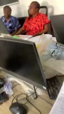The video shows an office scene with two women sitting at a desk covered with computers and office supplies. The woman on the right is wearing a red and orange patterned shirt, while the woman on the left is wearing a blue patterned top. They appear to be engaged in a conversation. The desk is cluttered with a computer monitor, mouse, and various papers. In the background, other people are seen standing and moving around in a hallway or doorway. The overall impression is a busy office environment.