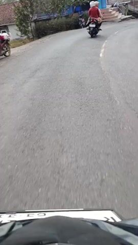 A first-person perspective video shows a motorcycle traveling down a paved road. The motorcycle is following another motorcycle carrying two people. The person in front of the lead motorcycle is wearing a red shirt and a white helmet. The second motorcycle is a dark color, and the rider is wearing a white helmet and a light-colored head covering. The road has a yellow line down the center. On the left side of the road, there is a building with blue and white paint and stairs. Further down the road, there are trees and houses. On the right side of the road, there are also trees and houses. Indonesian flags are visible on poles near some of the houses. The road appears to be in a rural or semi-rural area. The video is grainy and appears to be recorded on a mobile device.