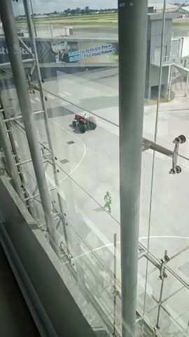A window view from inside an airport terminal shows a red tractor driving slowly across the tarmac. A ground worker wearing a neon vest is seen walking and then climbing on scaffolding. An advertisement for "Luxury service from takeoff to touchdown" is visible on the side of the building across the tarmac.