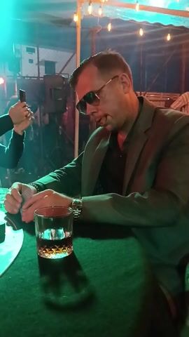 Here's a detailed description of the video:

The video shows a group of men playing poker at a table. The table is green and marked for "Texas Hold'em."  A man in a suit and sunglasses sits at the table, holding cards and with a glass of amber liquid nearby. Another man is taking a photo with his phone. The table is covered with poker chips and cards. The background includes a crowd of people, some holding flaming torches. The lighting is colorful and festive. The atmosphere appears to be lively and social.