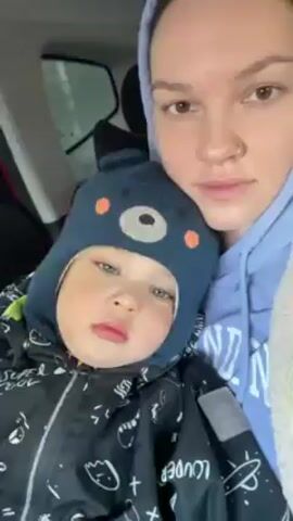 A woman wearing a blue hoodie and a nose ring is holding a young child wearing a black and white patterned snowsuit and a blue bear hat. They are inside a car, and the woman is taking a selfie. The child appears sleepy and is looking at the camera. The woman has a serious expression. The background shows the car's interior and a glimpse of a window with rain or snow on it.