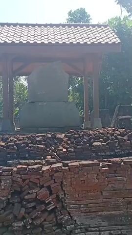 The video shows an outdoor archaeological site with ruins made of red bricks. In the foreground, there are piles of broken bricks, some forming low walls. In the midground, a covered structure with a tiled roof is supported by wooden posts. A large stone block is visible under the shelter. The background features lush green trees and a bright blue sky with some clouds. Further in the background, there are buildings, including houses with tiled roofs, and a dirt path. A motorcycle is parked near one of the buildings. Informational signs are posted on a fence in the background. The overall impression is one of an ancient site that is being preserved or excavated.