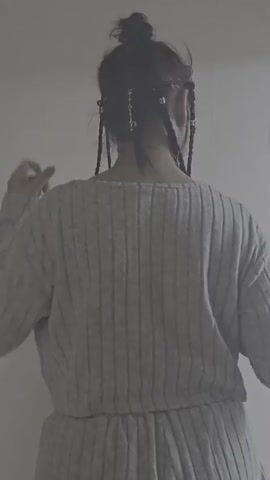 The video shows the back of a person with their hair styled in braids adorned with small silver charms. The person is wearing a grey ribbed sweater and grey pants. Their hands are positioned near their head, as if adjusting their hair or contemplating. The background is a plain, light-colored wall. The person's hair is partially pulled up into a bun, with several braids hanging down their back.