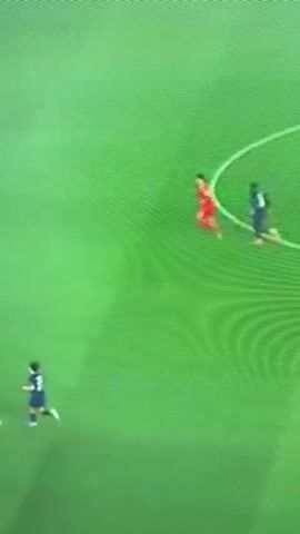 The video displays a football match from an elevated, slightly blurry perspective. The scene is set on a green football field, possibly illuminated by artificial lights, suggesting an evening or night game.

At the beginning of the clip, two players are visible in the mid-right of the frame, running side-by-side. One player is wearing a red kit, and the other is in a dark blue or black kit. A third player, also in a dark blue/black kit, is further away in the upper-left portion of the screen.

As the action unfolds, the player in the red kit makes contact with the player in the dark blue/black kit. The red-clad player then executes a tackle, bringing down the dark blue/black player, who falls to the ground. A football is briefly visible near their feet during the tackle. The player in red stands over the fallen player for a moment before moving away, while the tackled player remains on the ground. The third player continues to be visible in the distance, observing the play.

The video captures a dynamic moment of physical engagement during a football game. No text is visible in the frame. The provided context indicates the location is Lucerne, Switzerland.